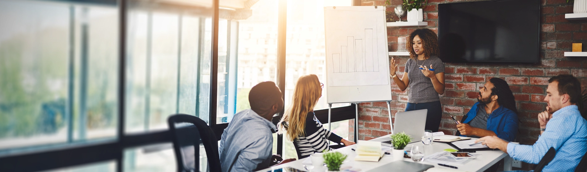 Woman presenting at a white board with a group of colleagues sitting around a meeting table in a conference room