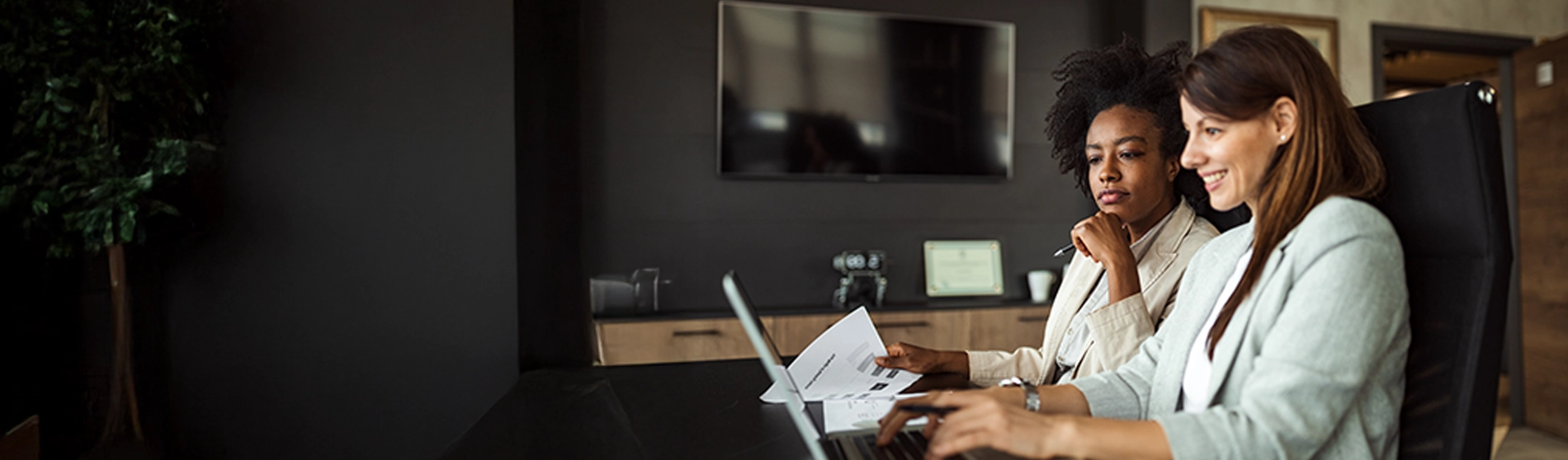 Two women sitting at desk looking over paperwork and a laptop