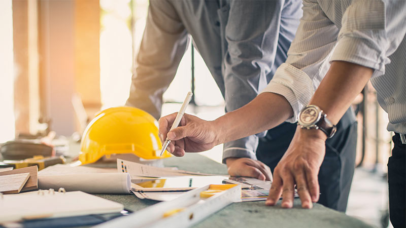 Close up of two people working at a construction desk full of tools, blueprints and a yellow hard hat