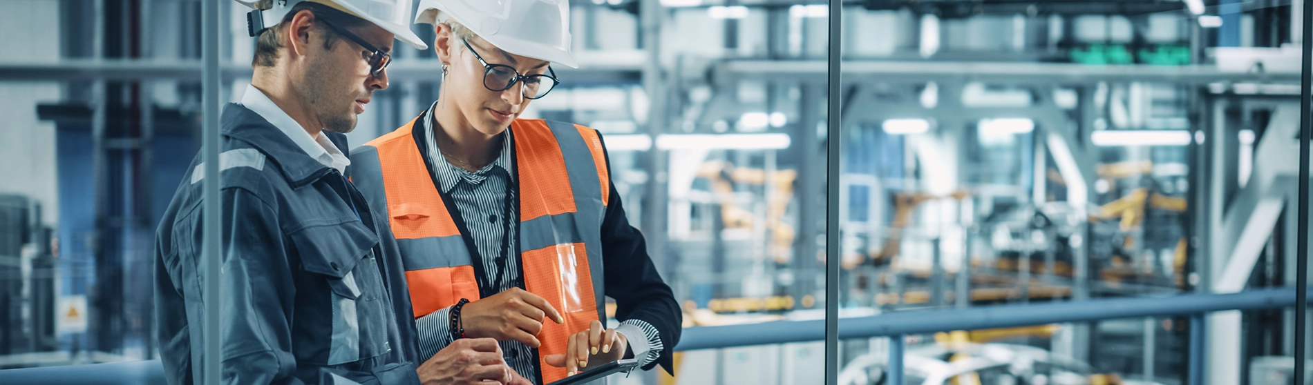 Two engineers in hard hats and safety vests reviewing information on a tablet in an industrial setting, surrounded by machinery and equipment.