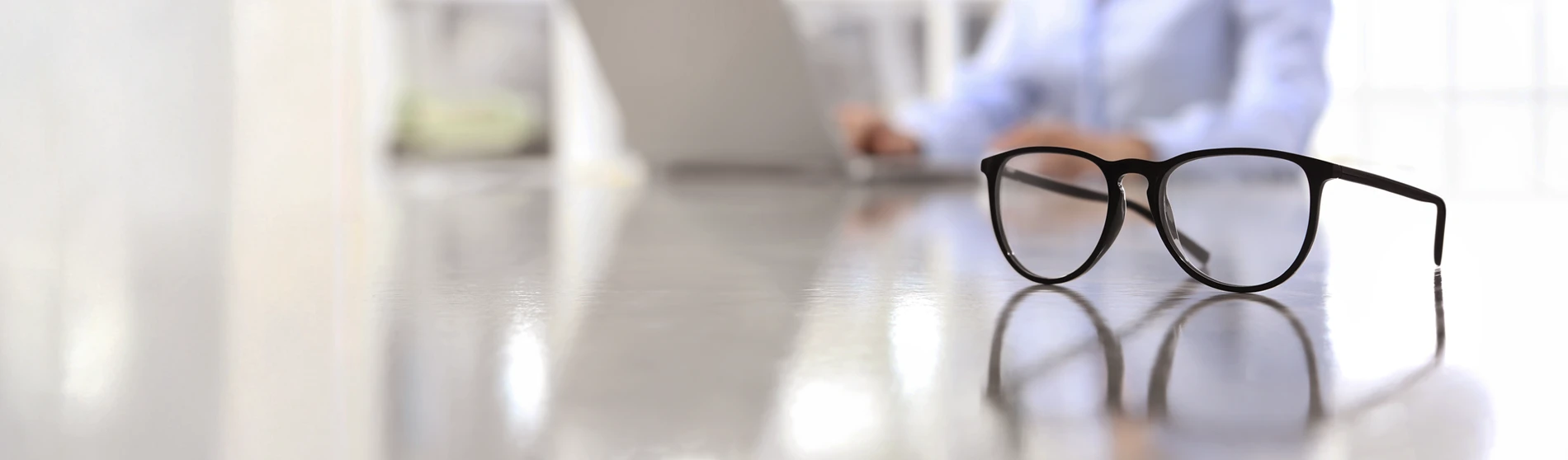 black framed glasses on a desk with woman on laptop in background