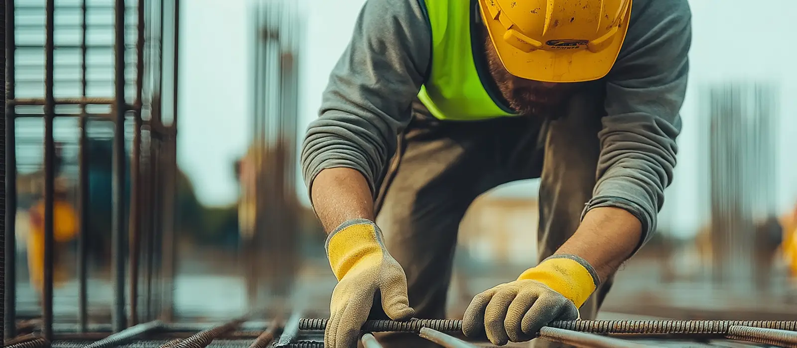 A construction worker in a yellow hard hat and reflective vest is kneeling on a construction site.