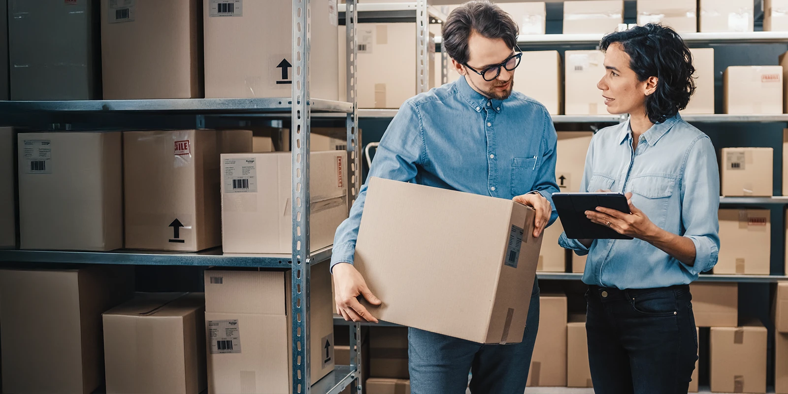 man holding box speaking to woman with tablet in storage room