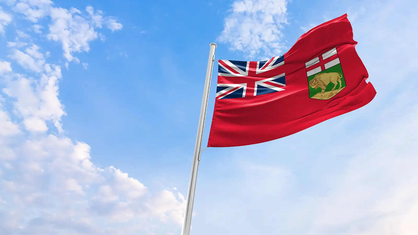 Un drapeau rouge avec l'Union Jack dans le coin supérieur gauche et un bouclier représentant un ours sur un fond vert dans la moitié droite, flottant sur un mât blanc contre un ciel bleu avec des nuages épars.