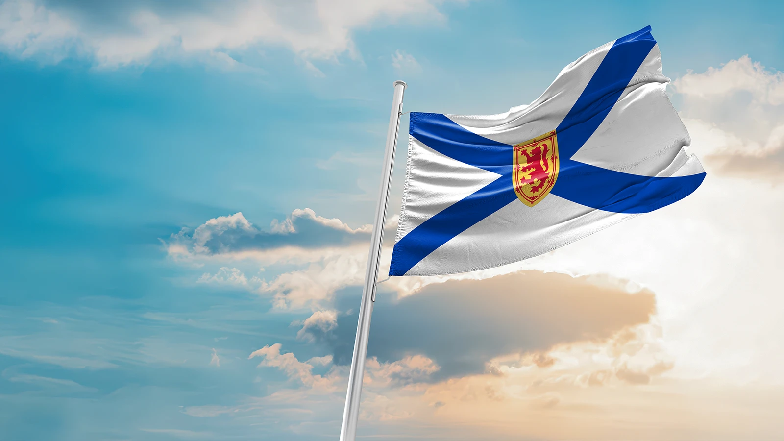 The image shows the flag of Nova Scotia, Canada, waving against a backdrop of a partly cloudy sky with the sun setting or rising. The flag features a blue saltire on a white field with the royal arms of Scotland in the center.